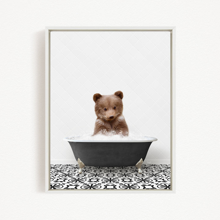 A cute brown bear cub sitting in a black bathtub filled with bubbles, looking directly at the camera.