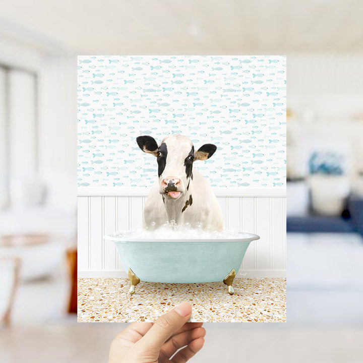 A hand holds up a framed image of a cow sitting in a bathtub filled with bubbles, surrounded by a pattern of fish.