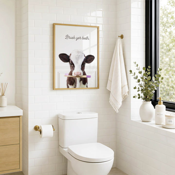 A bathroom with a white toilet, a framed poster of a cow holding a toothbrush, and a window with greenery outside.