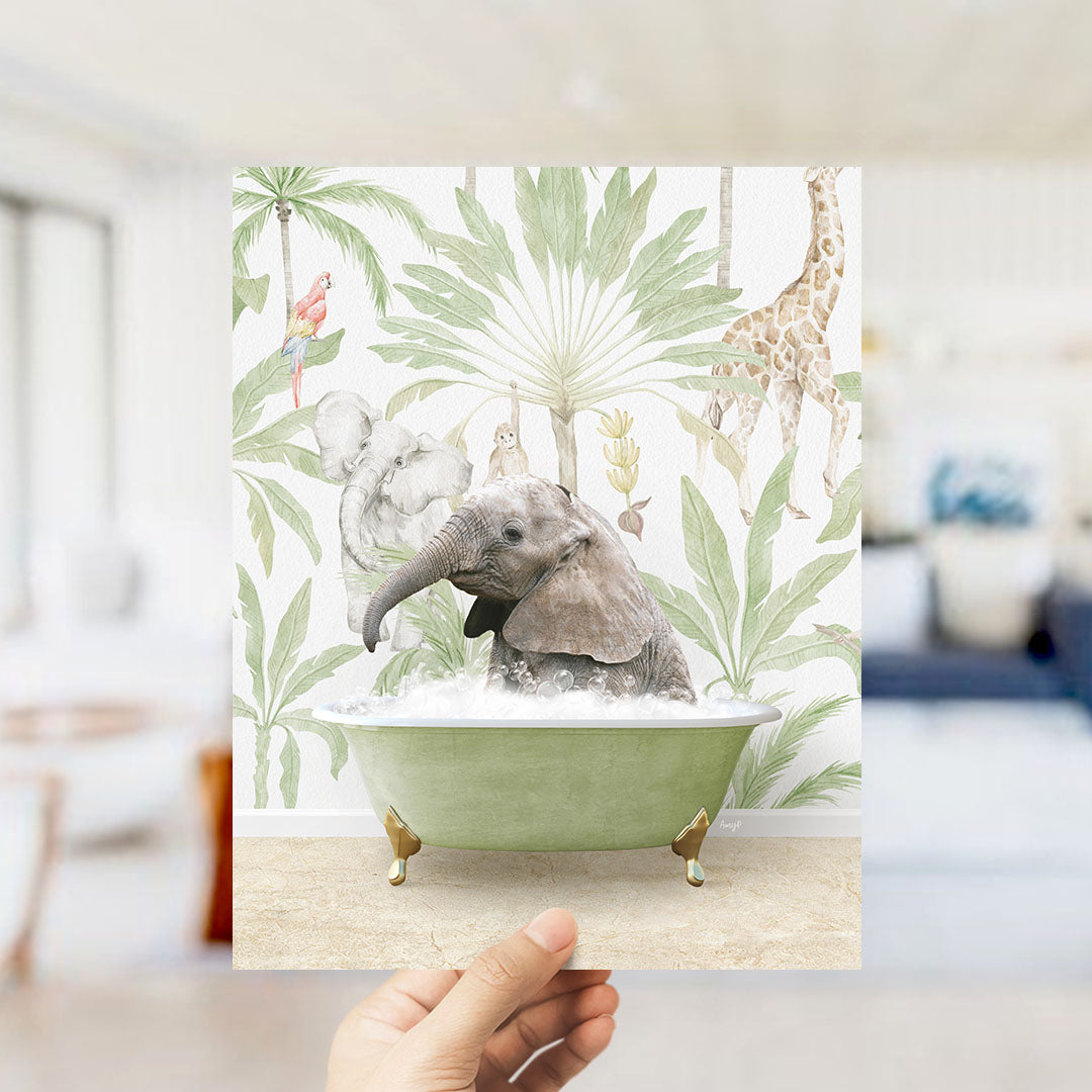 A hand holds up a framed print featuring an elephant sitting in a bathtub surrounded by tropical foliage, including a giraffe and palm trees.