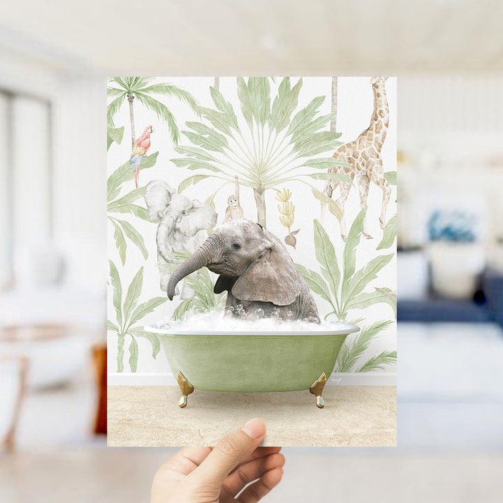 A hand holds up a framed print featuring an elephant sitting in a bathtub surrounded by tropical foliage, including a giraffe and palm trees.