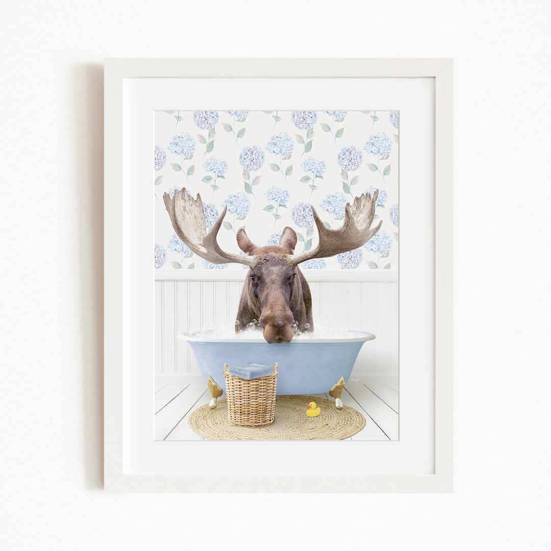 A moose with large antlers is sitting in a blue bathtub, surrounded by a floral patterned wall and a basket of rubber ducks.