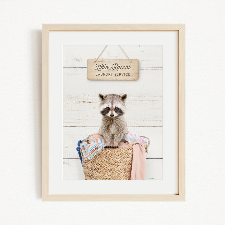 A raccoon sitting in a woven laundry basket, with a sign above that reads "Little Racal Laundry Service".