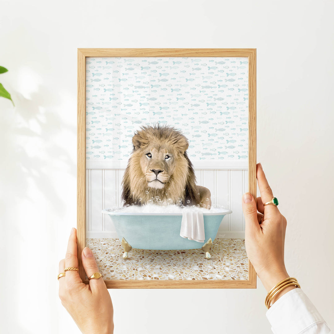 A person's hands are holding a framed image of a lion sitting in a bathtub filled with bubbles, with a towel draped over the side.