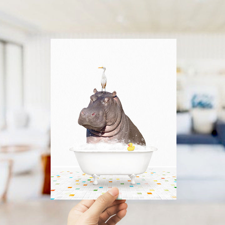 A hand holds up a framed image depicting a hippopotamus sitting in a bathtub, with a seagull perched on its head.