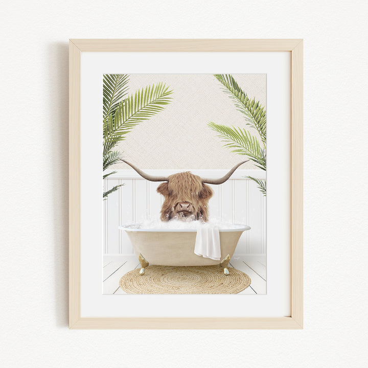 A framed image depicting a Highland cow with long horns sitting in a bathtub filled with bubbles, surrounded by palm fronds.