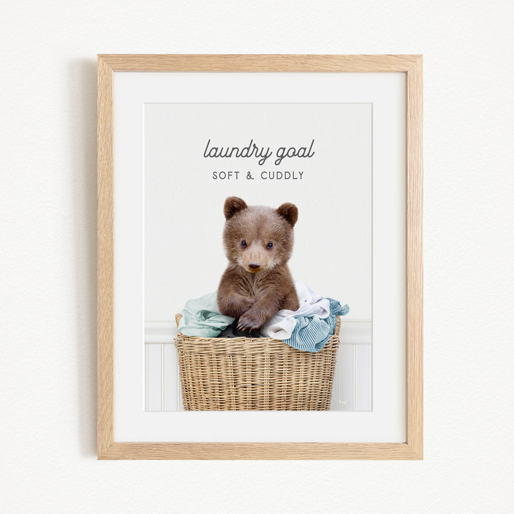 A cute brown bear sitting in a wicker laundry basket filled with clothes, with the text "laundry goal SOFT & CUDDLY" displayed above the bear.