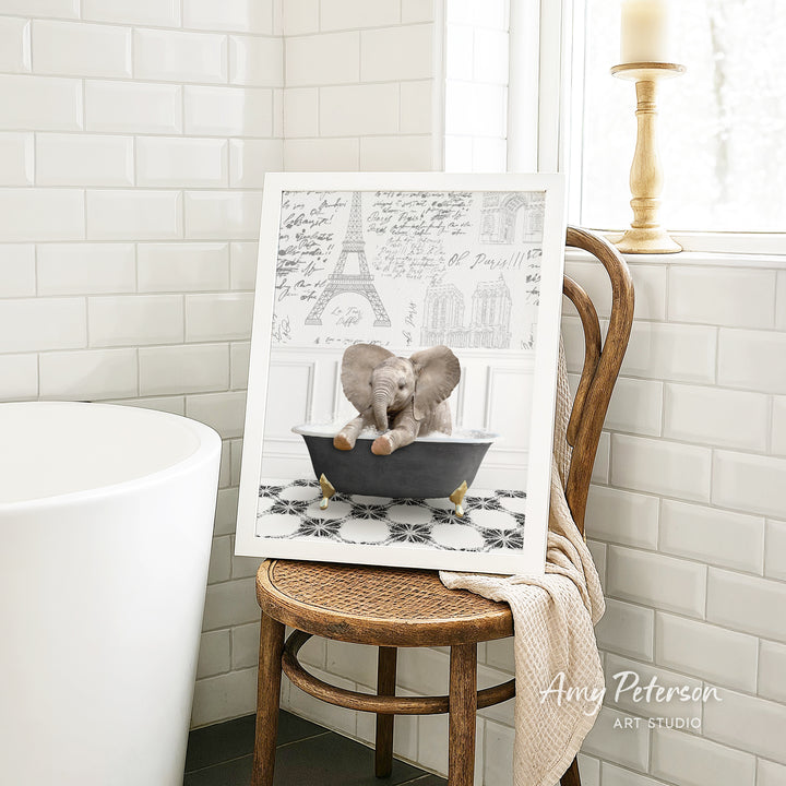 A framed image of a baby elephant sitting in a bathtub, with a Paris-themed background, is displayed on a wooden chair in a bathroom setting.