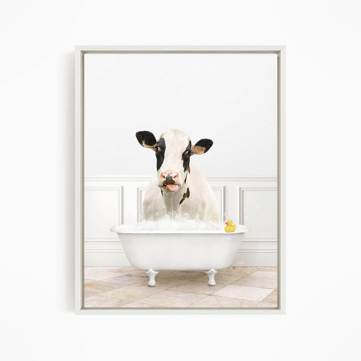 A black and white cow is sitting in a white bathtub filled with water, with a yellow rubber duck floating in the tub.