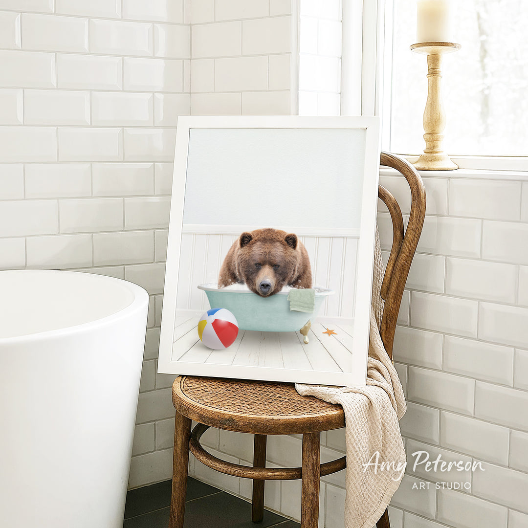 A framed image of a brown bear sitting in a bathtub, surrounded by a beach ball and a starfish, is displayed on a wooden chair in a bathroom setting.