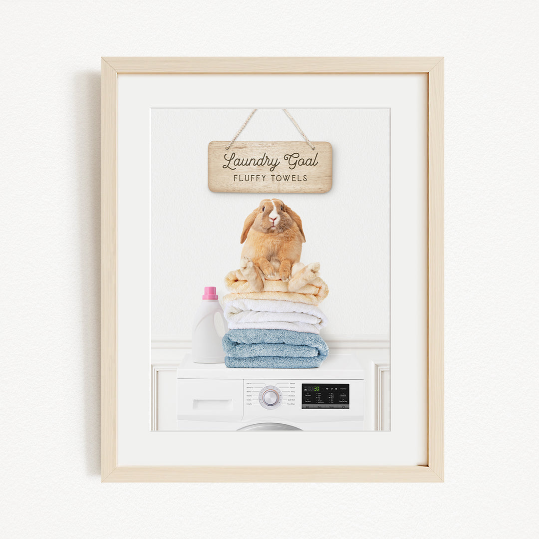 A framed image of a rabbit sitting on a stack of folded towels, with a sign above that says "Laundry goal Fluffy towels".