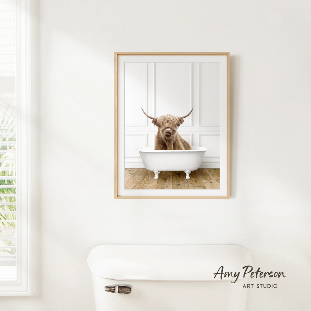 A framed image of a Highland cow sitting in a bathtub is hanging on a white wall in a bathroom.