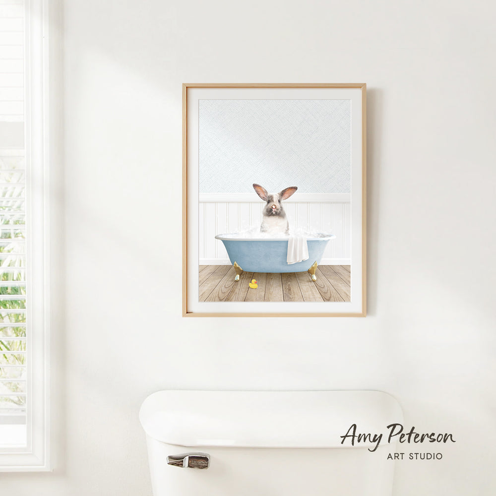 A framed image of a rabbit sitting in a bathtub, with a towel draped over its body, is hanging on a white wall.