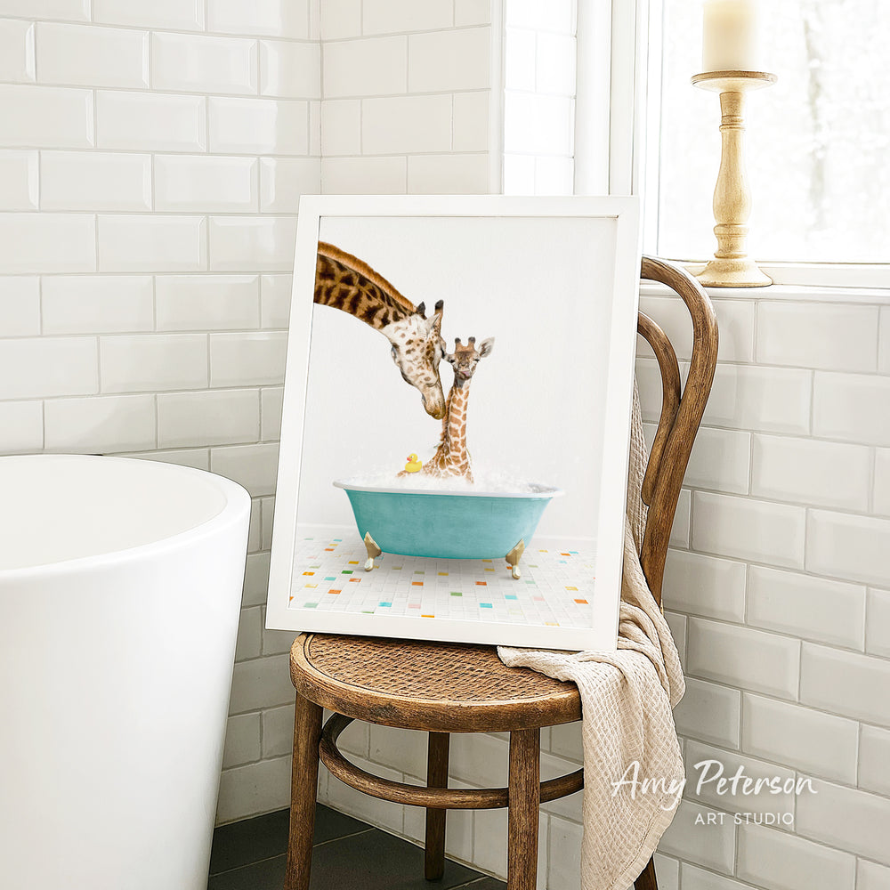 A framed image of a giraffe and a baby giraffe sitting in a blue bathtub, with a towel draped over the back of a wooden chair in a bathroom setting.