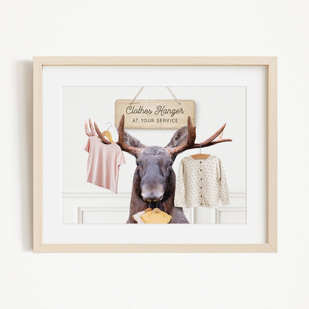 A framed image depicting a moose with antlers holding clothes hangers, with a sign that reads "Clothes Hanger at your Service".