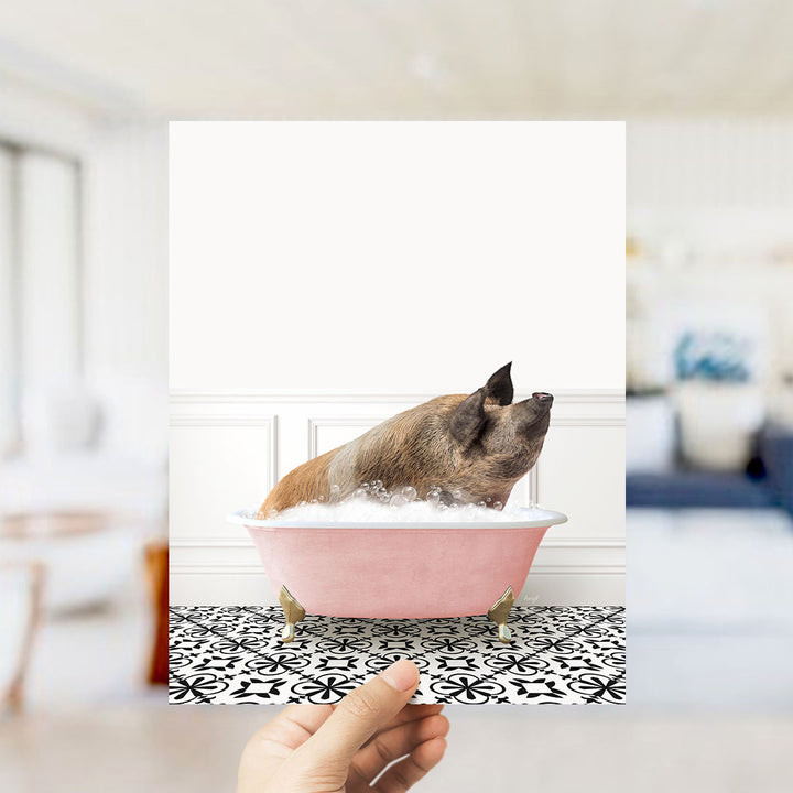 A hand holds up a framed image of a pig sitting in a pink bathtub filled with bubbles, surrounded by a black and white patterned floor.