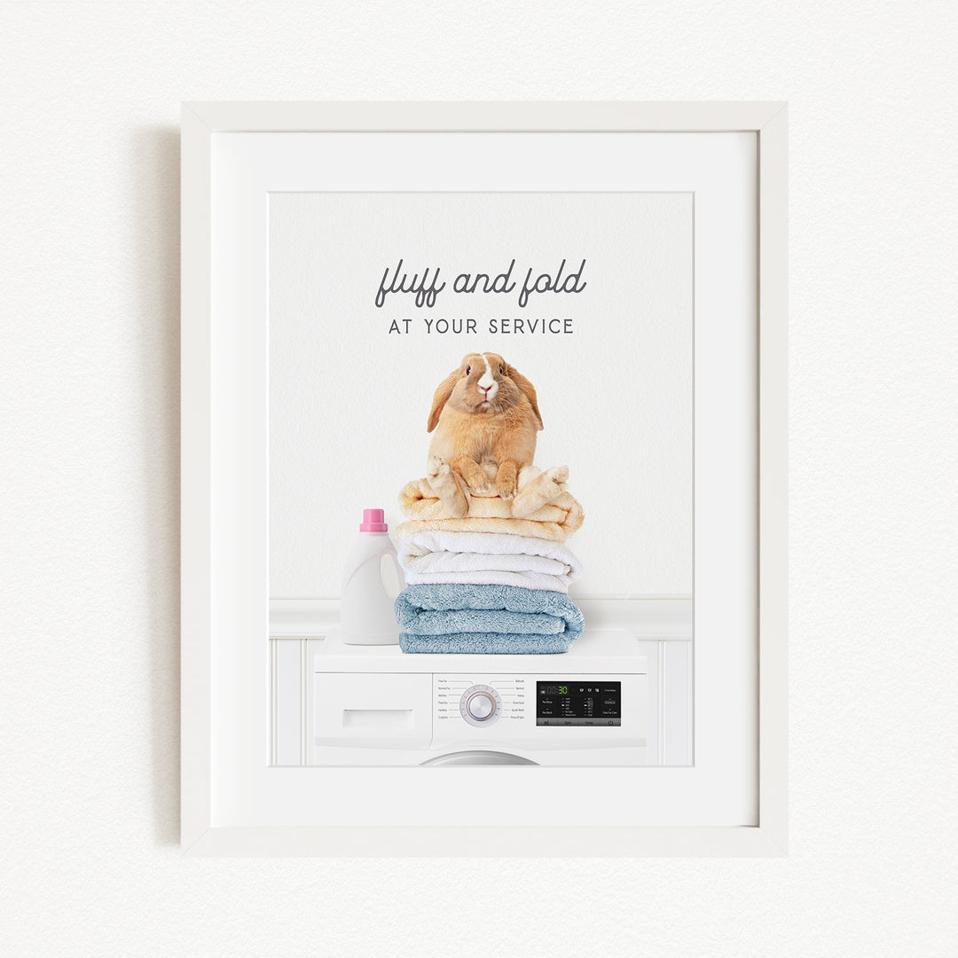 A framed image of a fluffy rabbit sitting atop a stack of folded towels, with the text "fluff and fold at your service" written above the rabbit.