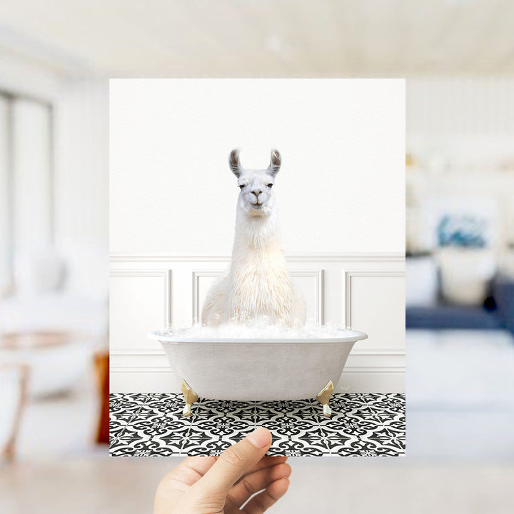 A white llama sitting in a white bathtub filled with bubbles, with a hand holding the image up to the viewer.