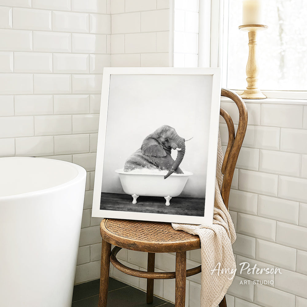 A framed black and white photograph of a baby elephant sitting in a bathtub, with a towel draped over the back of a wooden chair in the foreground.