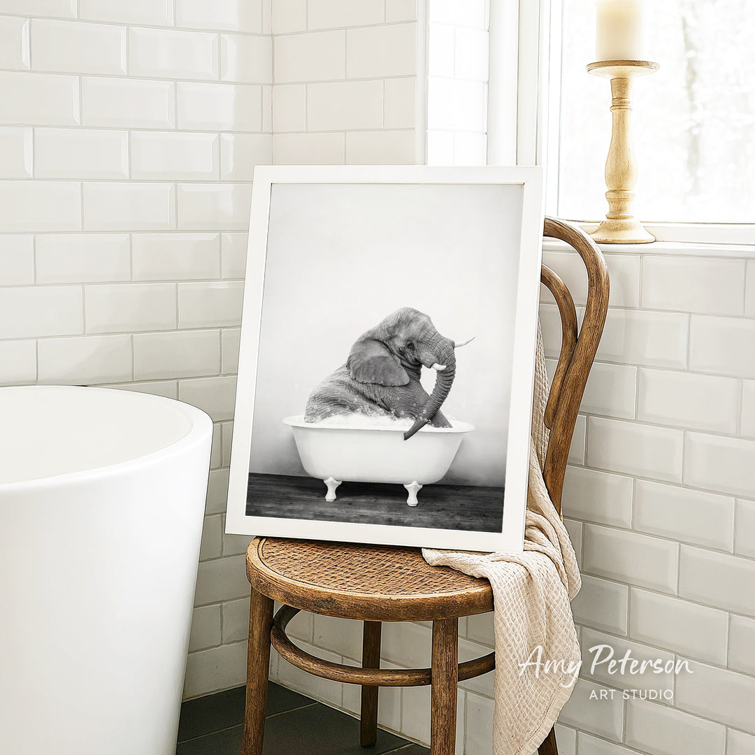 A framed black and white photograph of a baby elephant sitting in a bathtub, with a towel draped over the back of a wooden chair in the foreground.