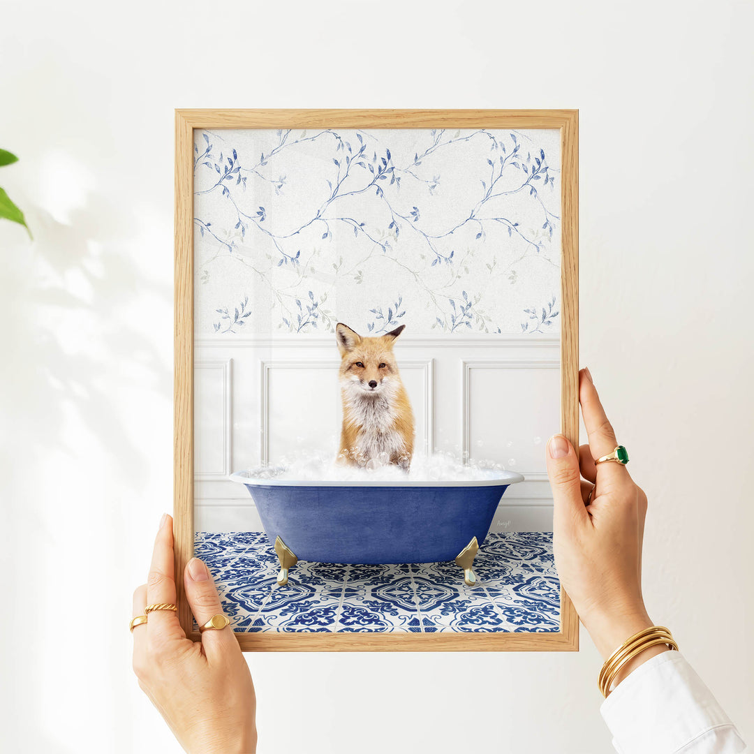 A person's hands are holding a framed picture of a fox sitting in a blue bathtub filled with water, surrounded by a blue and white floral pattern.