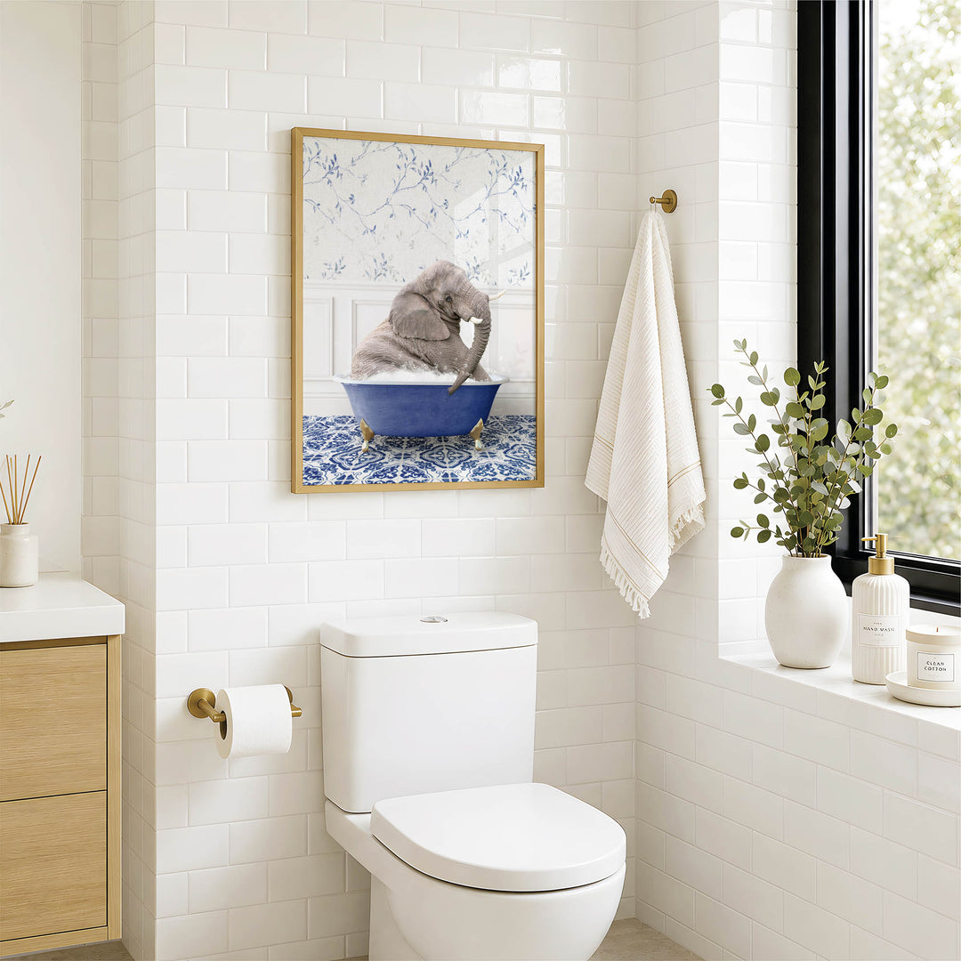 A bathroom with a white toilet, a framed picture of an elephant sitting in a blue bathtub, and a window with greenery outside.