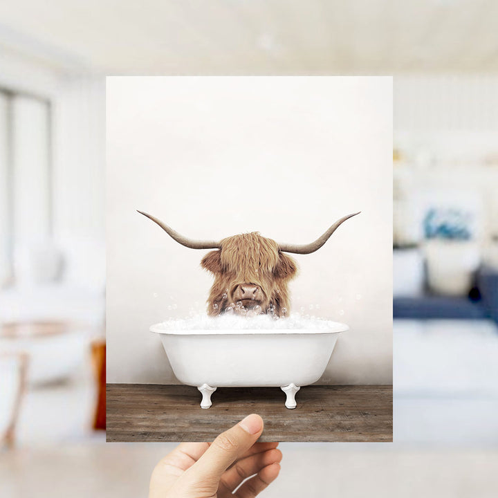 A Highland cow with large horns is sitting in a white bathtub filled with bubbles, as a person holds up the image.