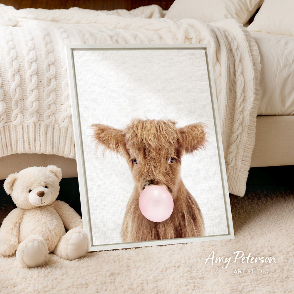 A framed image of a brown cow blowing a bubble gum bubble, accompanied by a teddy bear on a bed.