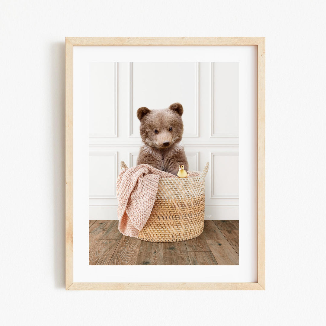 A brown bear sitting in a woven basket, with a pink blanket and a small toy duck nearby.