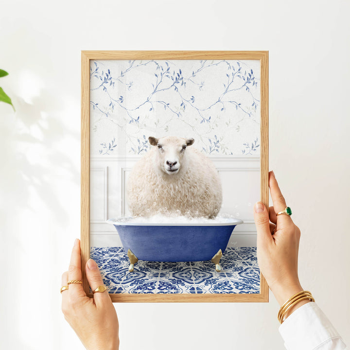 A person's hands are holding a framed picture of a white sheep sitting in a blue bathtub filled with bubbles, against a white wall with a blue floral pattern.