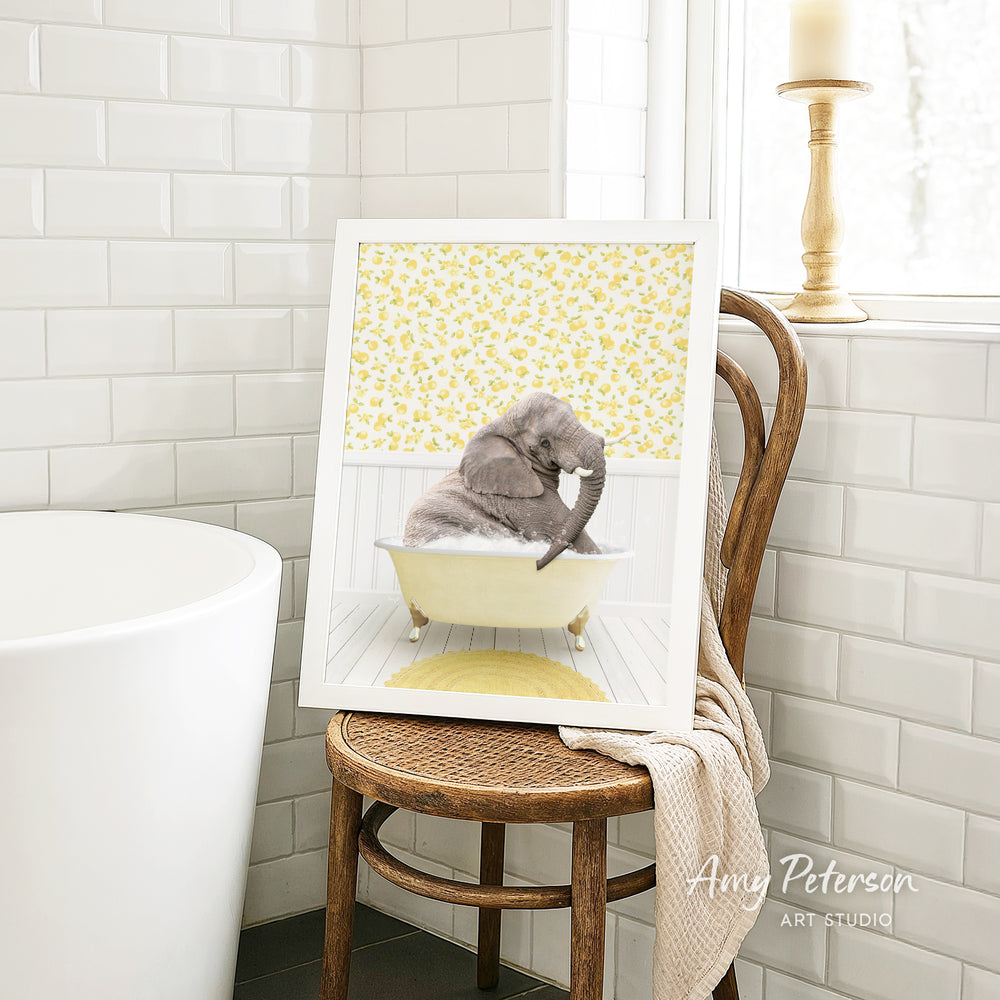 A framed image of a gray elephant sitting in a yellow bathtub, surrounded by a yellow floral pattern, is displayed on a wooden chair in a bathroom setting.