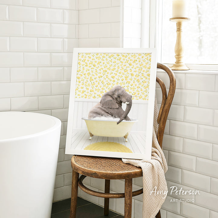 A framed image of a gray elephant sitting in a yellow bathtub, surrounded by a yellow floral pattern, is displayed on a wooden chair in a bathroom setting.