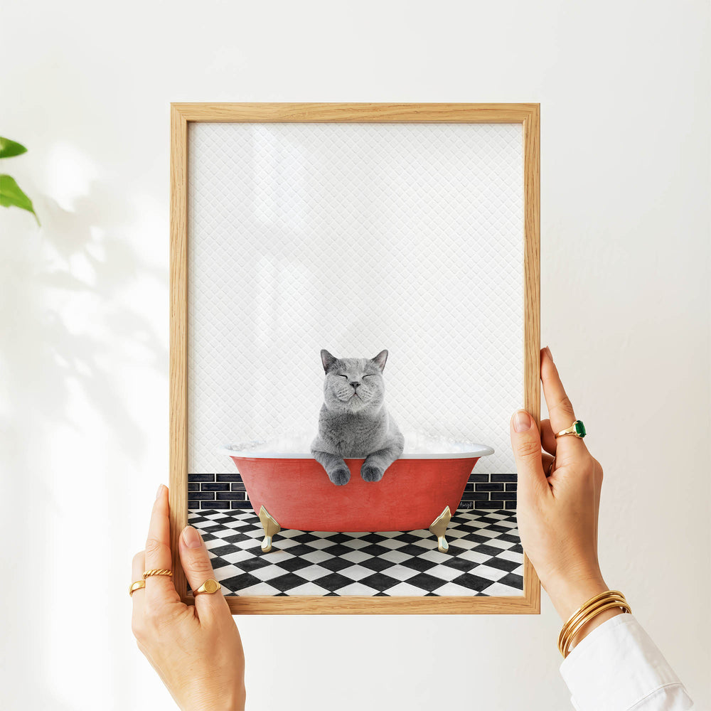 A person's hands are holding a framed picture of a gray cat sitting in a red bathtub filled with water, with a black and white checkered floor beneath it.