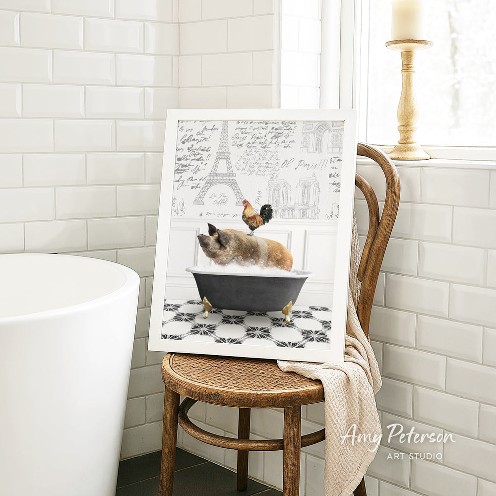 A framed image of a cat sitting in a bathtub, with a rooster perched on its head, is displayed on a wooden chair in a bathroom setting.