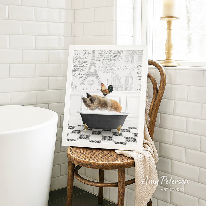 A framed image of a cat sitting in a bathtub, with a rooster perched on its head, is displayed on a wooden chair in a bathroom setting.