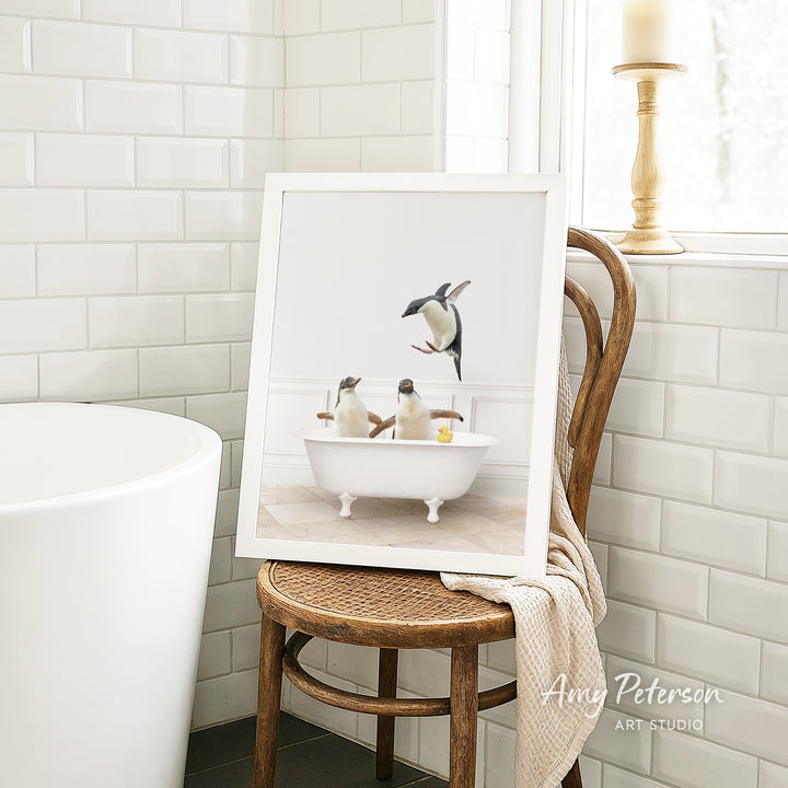 A framed image of two penguins sitting in a bathtub, with one penguin standing and the other sitting, is displayed on a wooden chair in a bathroom setting.