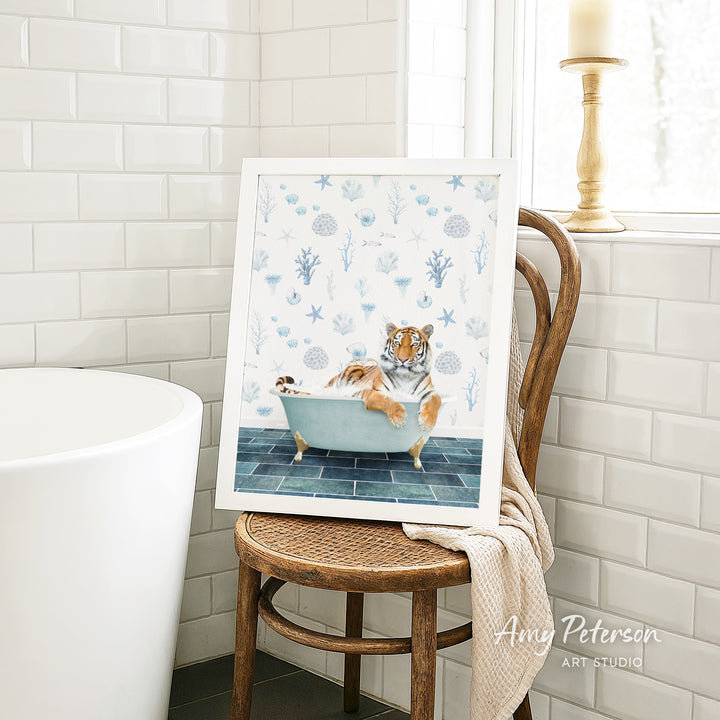 A framed painting of a tiger sitting in a bathtub, surrounded by blue and white floral patterns, is displayed on a wooden chair in a bathroom setting.