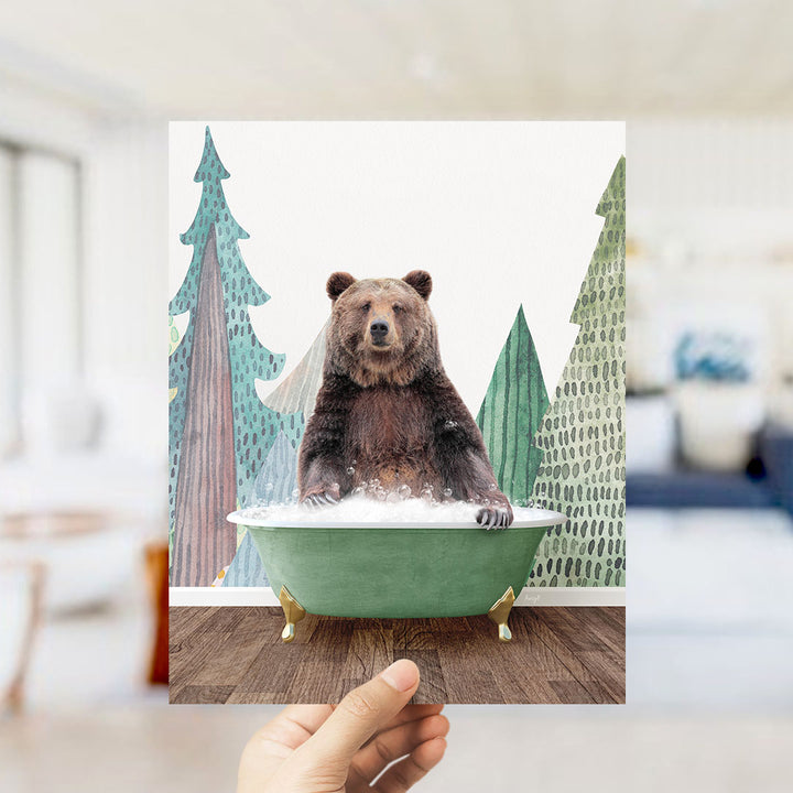 A hand is holding up a framed image of a brown bear sitting in a green bathtub, surrounded by a forest scene with trees and a blue sky.
