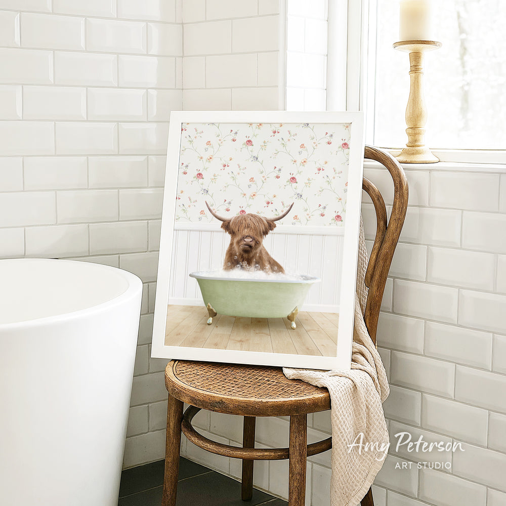 A framed image of a bull with horns sitting in a bathtub, surrounded by a floral pattern, is displayed on a wooden chair in a bathroom setting.