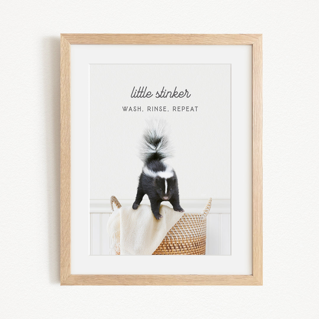A framed image of a black and white skunk sitting on a woven basket, with the text "little stinker" and "WASH. RINSE. REPEAT" written above it.