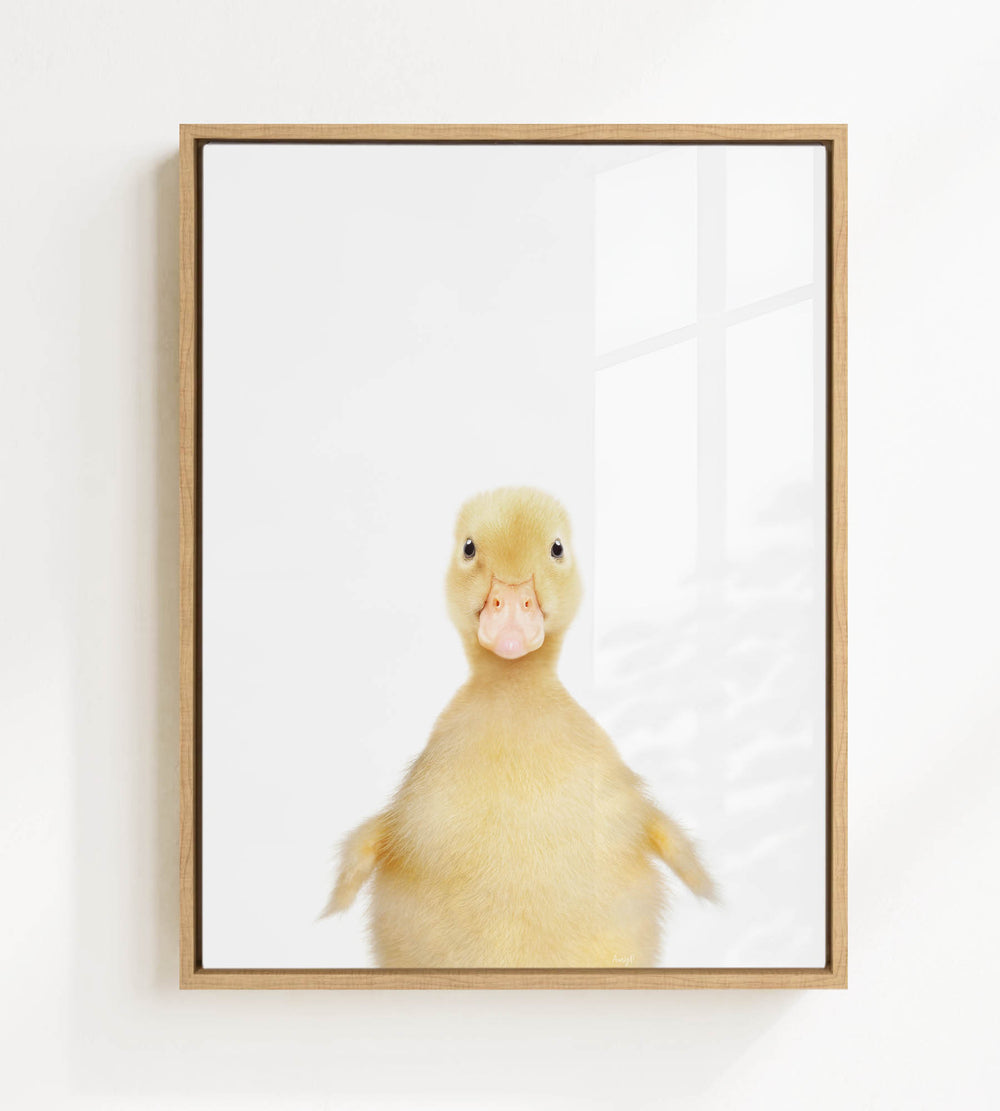 A cute yellow duck with a pink beak is the main subject of the framed print, which is displayed on a white wall.