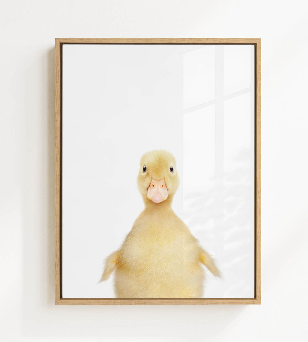 A cute yellow duck with a pink beak is the main subject of the framed print, which is displayed on a white wall.
