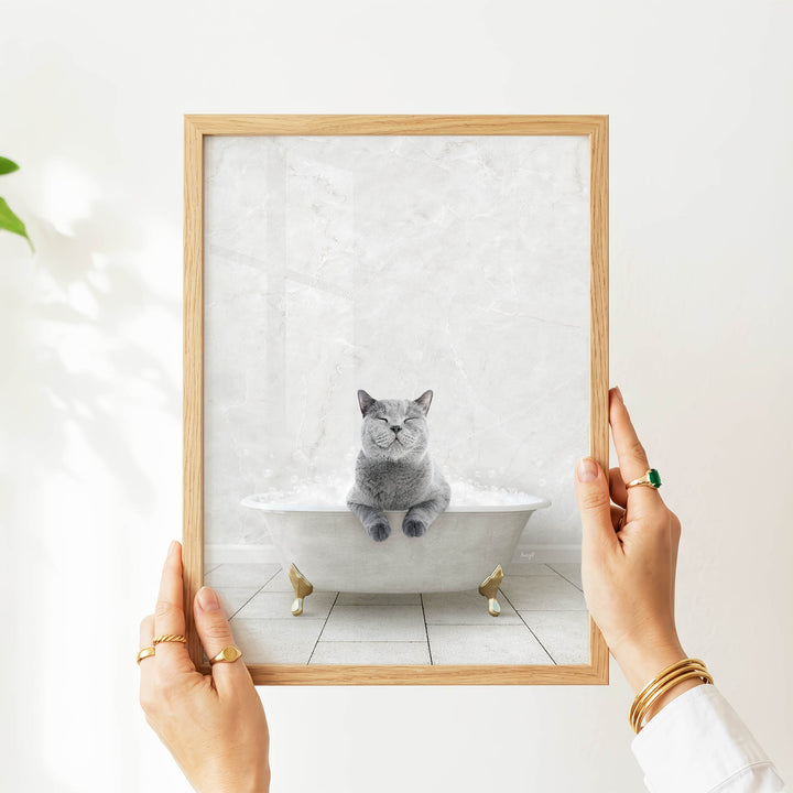 A person's hands are holding a framed picture of a gray cat sitting in a bathtub filled with water, with its eyes closed and appearing to be enjoying the bath.