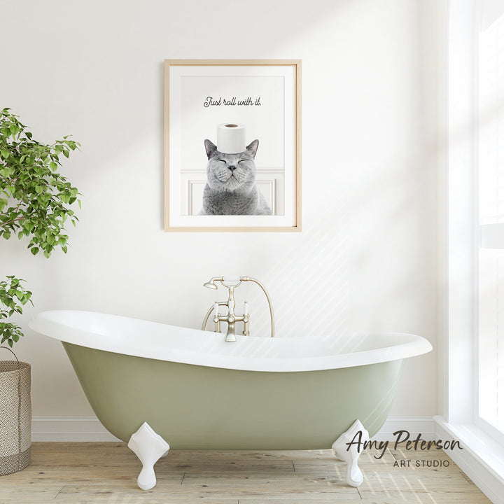 A framed image of a cat with a roll of toilet paper on its head is displayed on a white wall above a green clawfoot bathtub in a bright, minimalist bathroom.