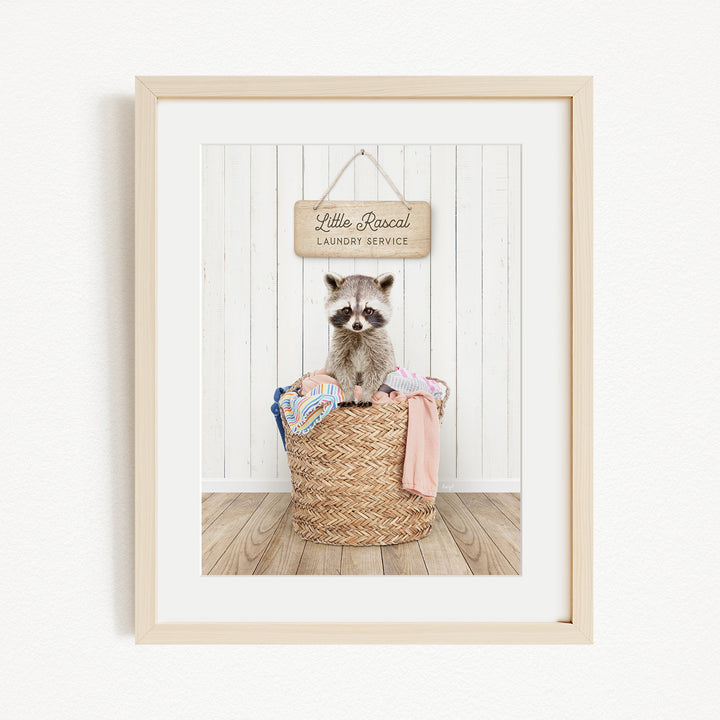 A raccoon sitting in a woven laundry basket, with a sign above that reads "Little Racquel Laundry Service".