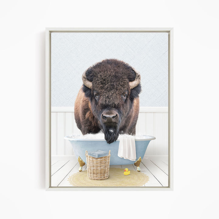 A buffalo with large horns is sitting in a blue bathtub filled with water, surrounded by a yellow rubber duck and a basket.