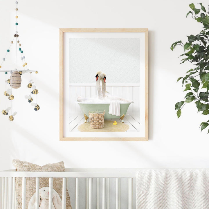 A framed image of a swan sitting in a bathtub, surrounded by a basket of bath toys, is displayed on a white wall in a nursery setting.