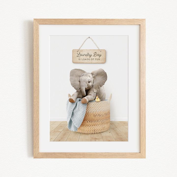 A framed image of a baby elephant sitting in a woven basket, with a sign above that says "Laundry Day is loads of fun".