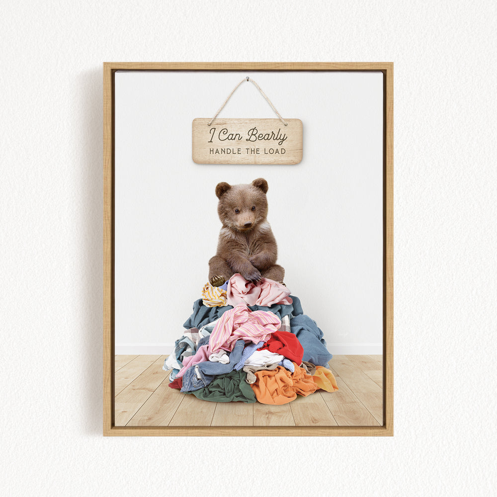 A teddy bear sitting atop a pile of clothes, with a wooden sign above that reads "I Can Bearly Handle the Load".