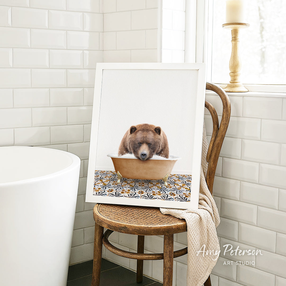 A framed image of a bear sitting in a bathtub, with a patterned towel draped over the back of a wooden chair in a bathroom setting.